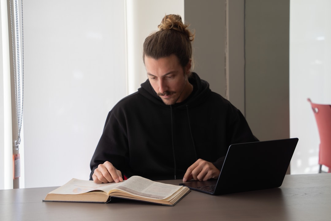 Man reads a book while using his laptop.
